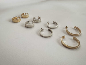 Classic gold and silver hoop earrings arranged in pairs, featuring thick band designs in both small and medium sizes on a light grey background