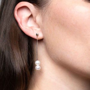 Close-up of a woman's ear adorned with a double pearl drop earring. The earring features two white pearls suspended from a slim silver rod with a stud backing, showcasing a modern and sophisticated look.