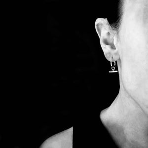 Close-up black and white image of a woman wearing minimalist small hoop earrings with a horizontal bar pendant, styled against a dark background.

